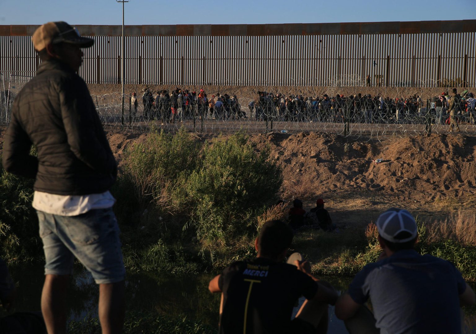 Fotografía de archivo de unos migrantes que esperan cerca de la puerta 42 del muro fronterizo en Ciudad Juárez, mientras integrantes de la Guardia Nacional del Estado de Texas, rodean con barricadas de púas la línea divisoria entre Ciudad Juárez y El Paso. EFE/Luis Torres