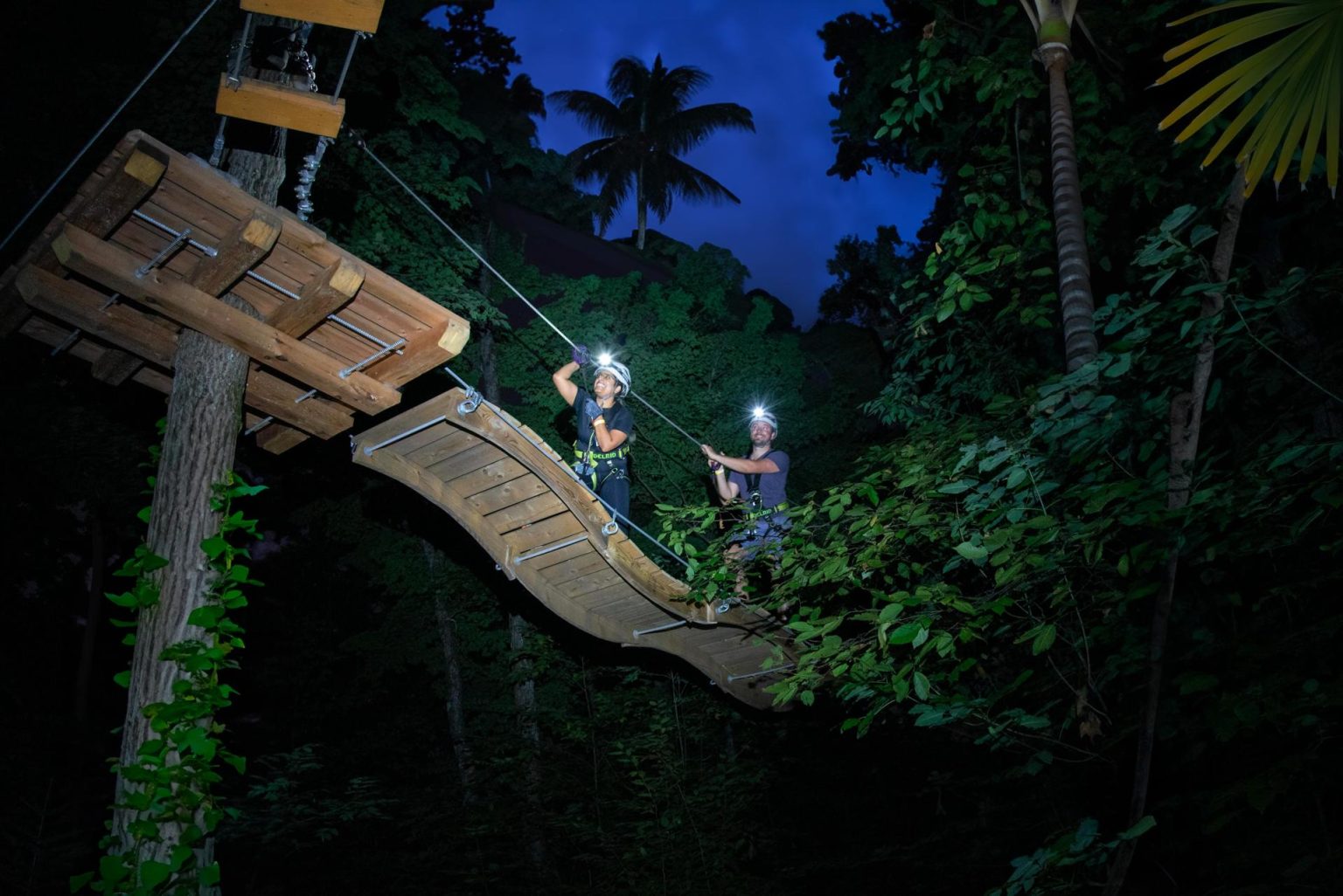 Fotografía cedida por The Trekking Group donde aparece dos personas mientras navegan entre árboles sobre tiras de madera durante su participación en la atracción nocturna 'Night Trek' ubicada en Jungle Island, el parque de ecoaventuras situado en la isla Watson de Miami, Florida. EFE/The Trekking Group /SOLO USO EDITORIAL /NO VENTAS /SOLO DISPONIBLE PARA ILUSTRAR LA NOTICIA QUE ACOMPAÑA /CRÉDITO OBLIGATORIO