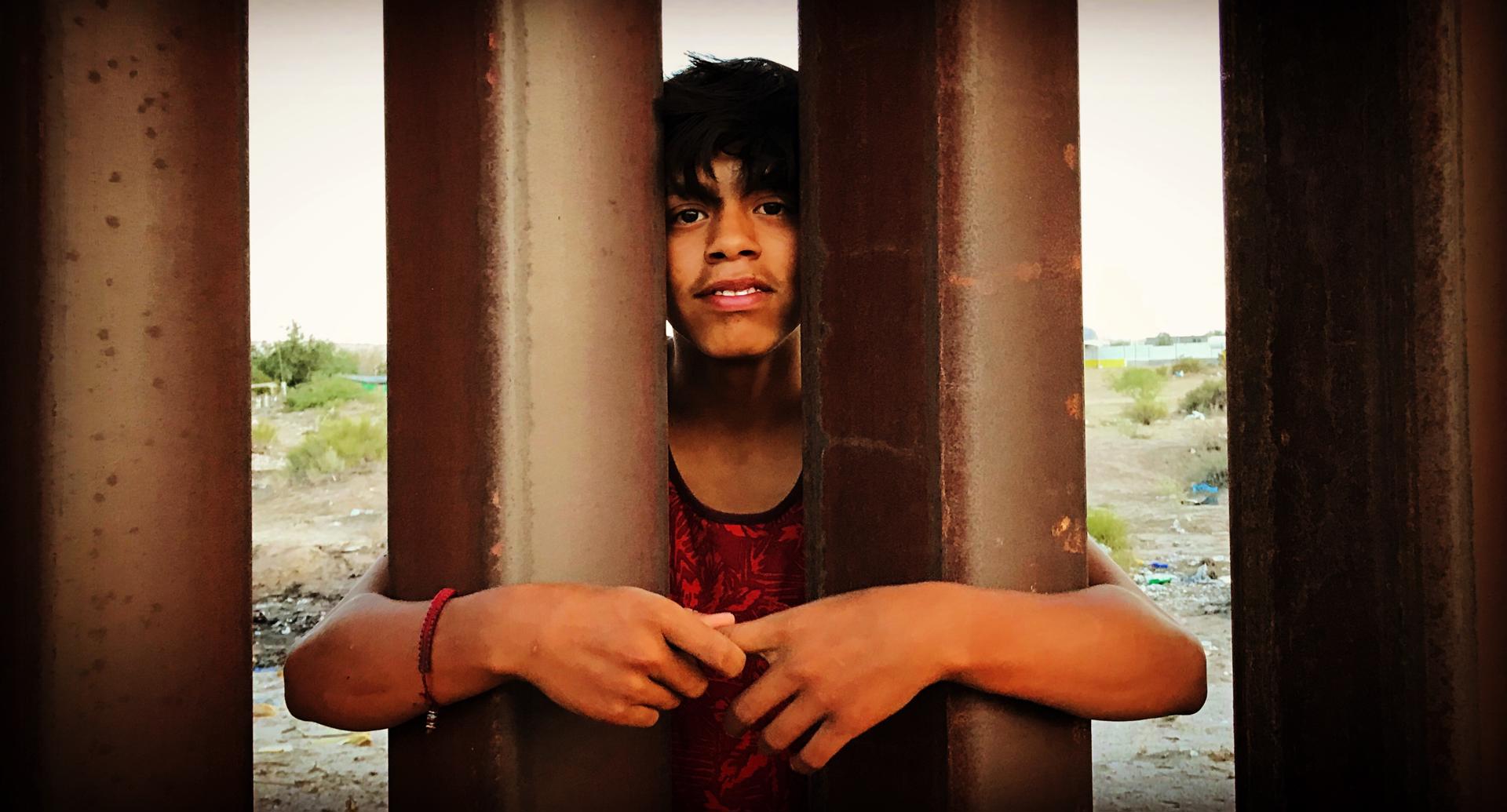 Fotograma cedido por Juan Hernández donde aparece un joven mexicano aferrado a la valla fronteriza, durante una escena del documental 'Frontera adentro' que habla sobre "el complejo industrial fronterizo", en referencia al uso de miles de millones de dólares en la militarización de la frontera y el establecimiento de docenas de centros de detención y uso de tecnología de punta. EFE/Juan Hernández /SOLO USO EDITORIAL /NO VENTAS /SOLO DISPONIBLE PARA ILUSTRAR LA NOTICIA QUE ACOMPAÑA /CRÉDITO OBLIGATORIO
