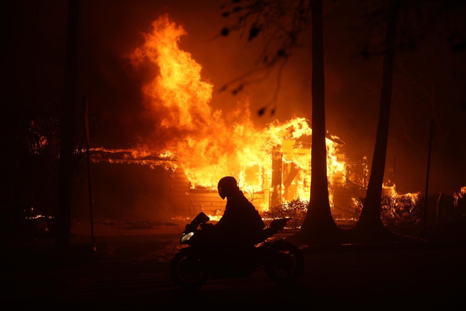 Una persona conduce su motocicleta junto a una casa en llamas durante los incendios en Palisades , California, Estados Unidos. EFE/ Allison Dinner